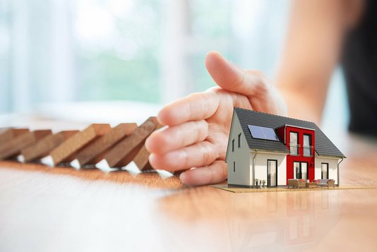 sicherheit-schutz-geborgenheit-familie-haus-geschuetzt-03 Schützende Hand vor einem Modellhaus.